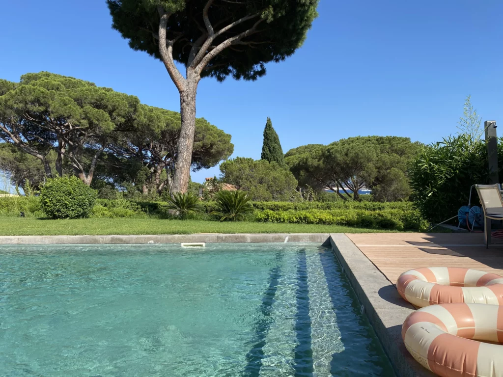 Vue d’un jardin avec piscine, végétation en arrière-plan et bouées gonflables au bord de l’eau, pour une ambiance estivale et conviviale.